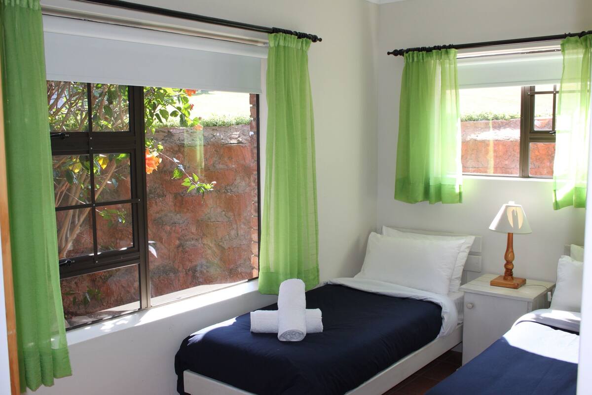 Una habitación de hotel con una cama, una mesita de noche con lámpara y una ventana con cortinas verdes.