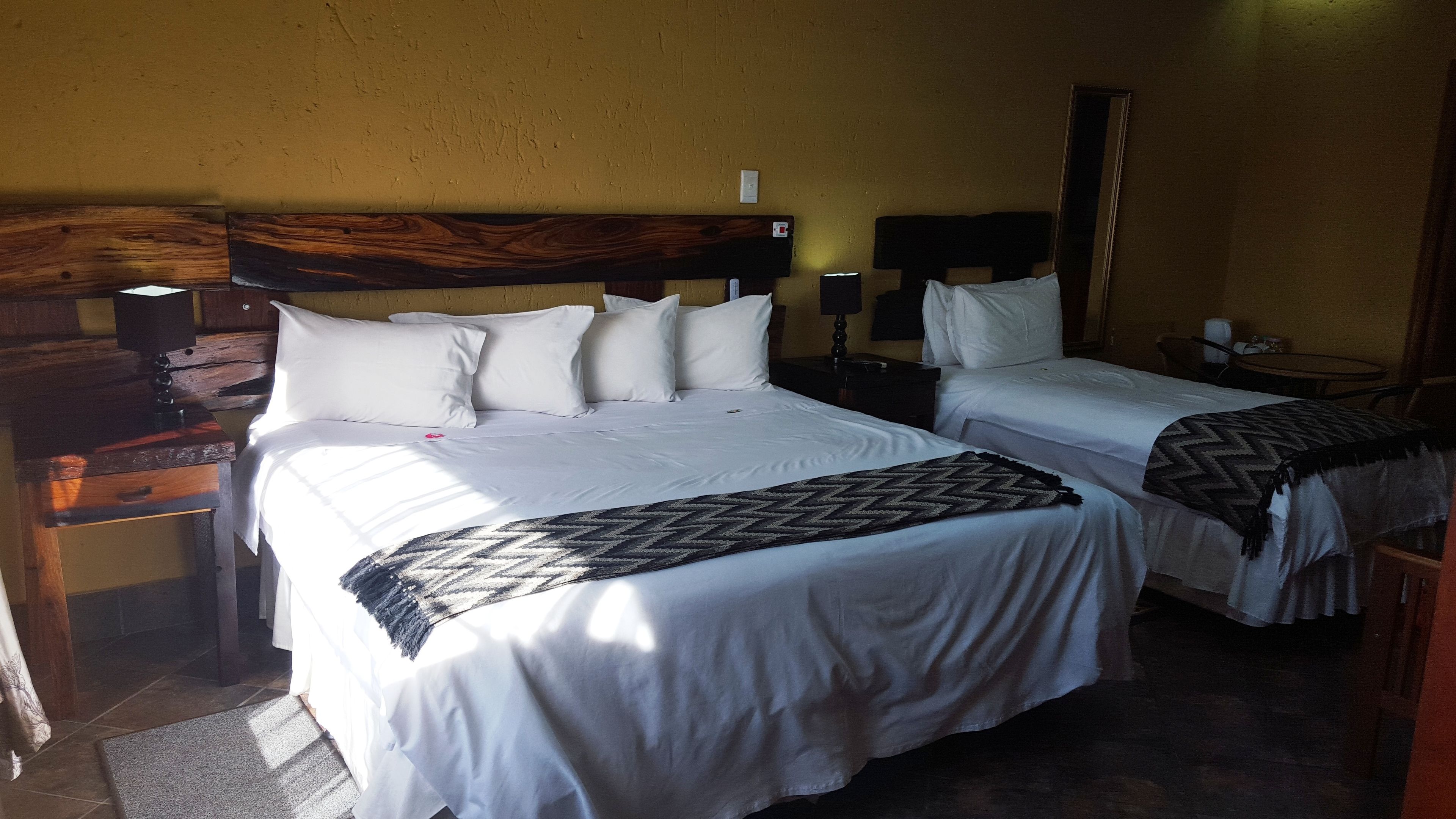 A hotel room with two beds, wooden headboards, white bedding, a patterned throw, a nightstand, a lamp, and a mirror.