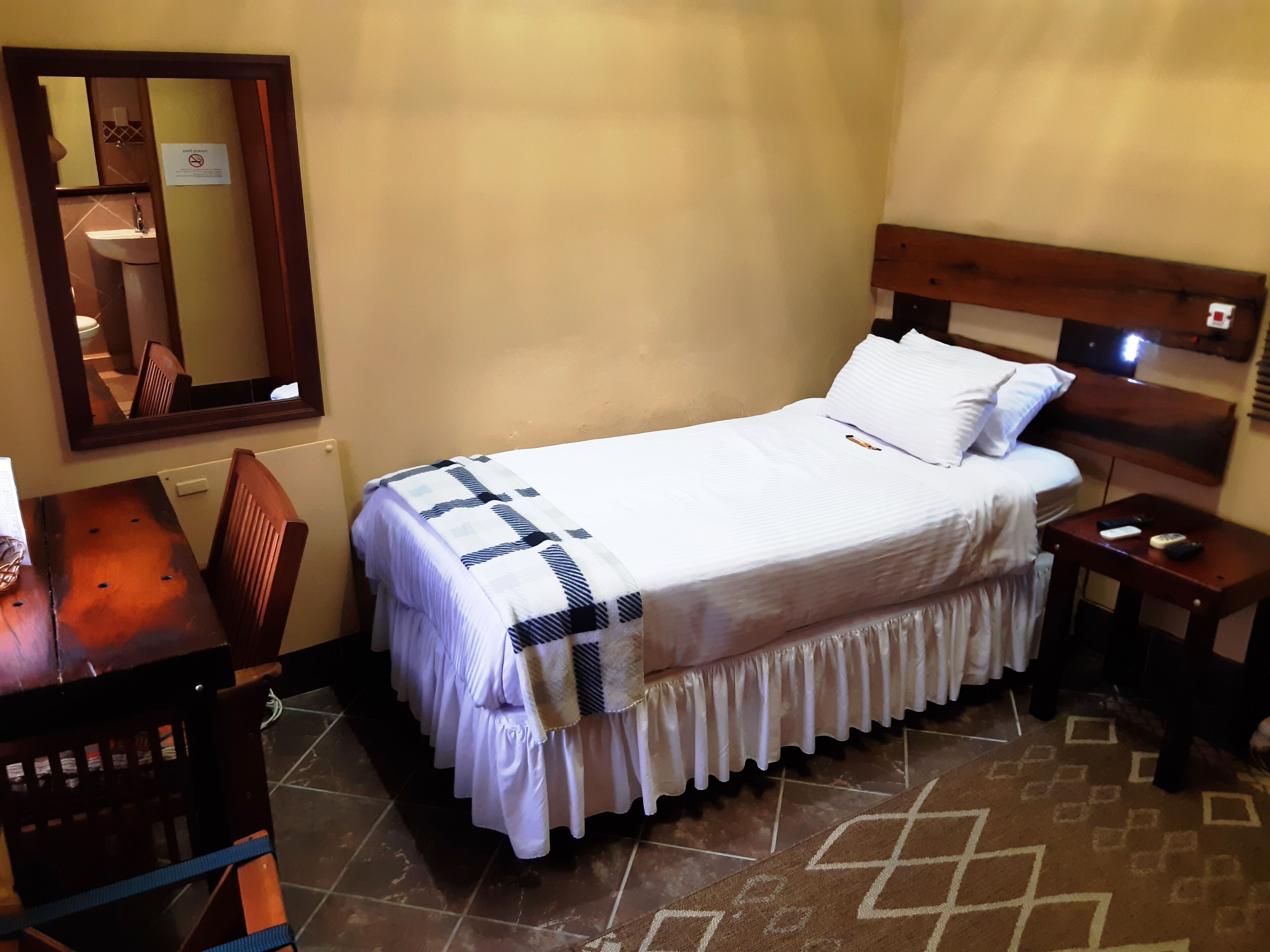 Una cama bien tendida con un edredón blanco y un borde decorativo, una mesita de noche de madera con un control remoto y un espejo en la pared.