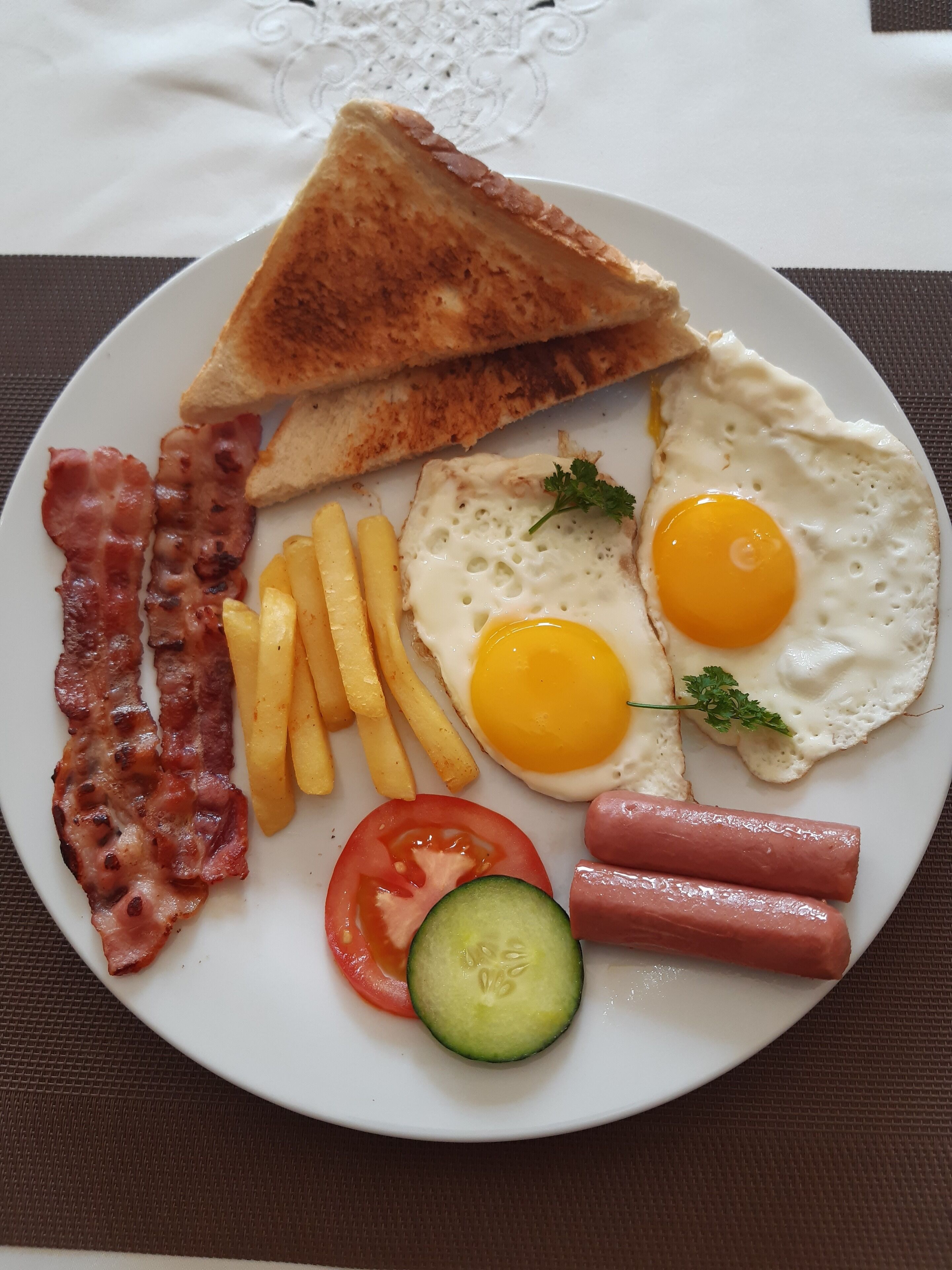 A plate of breakfast food including toast, eggs, bacon, sausages, fries, a tomato, and a cucumber slice.