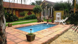 A swimming pool with a tiled deck, surrounded by greenery and a building with a red roof. - Acre of Africa Guest Venue (Boksburg)