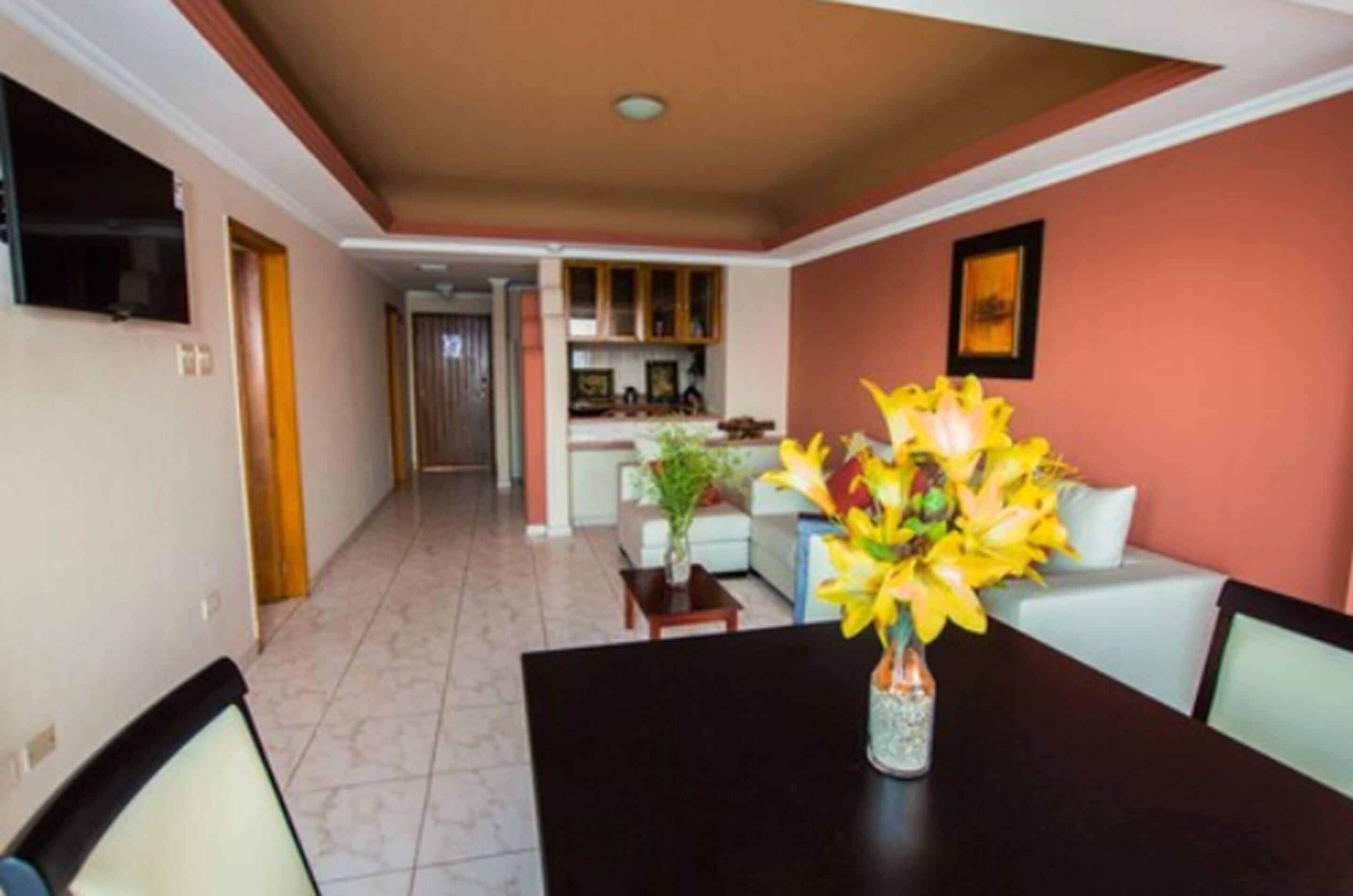 A dining area with a table and chairs, a vase with flowers, and a kitchen in the background.