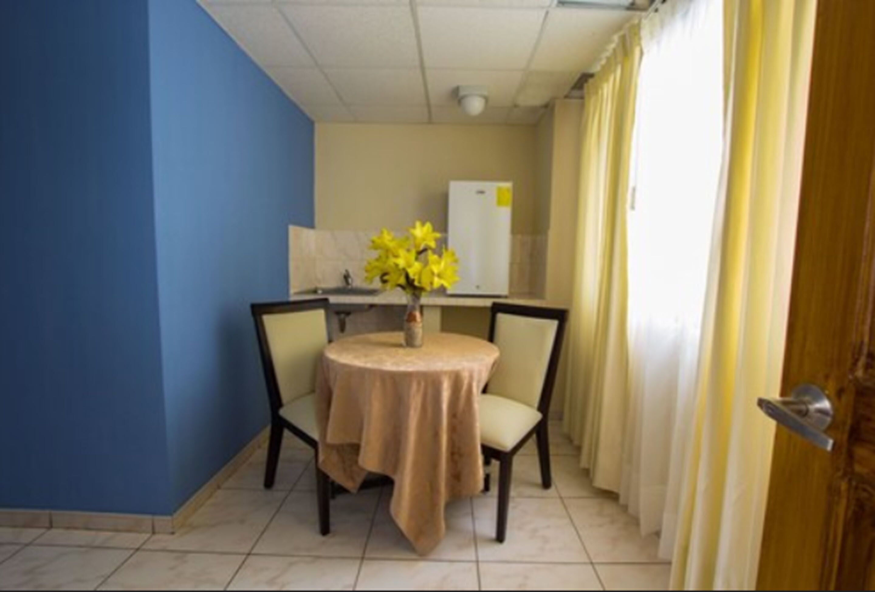 Een kleine kamer met een ronde tafel, twee stoelen, een vaas met gele bloemen, een witte koelkast en een deur met gordijn.