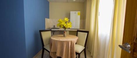 Una habitación pequeña con una mesa redonda, dos sillas, un florero con flores amarillas, un refrigerador blanco y una puerta con cortina.