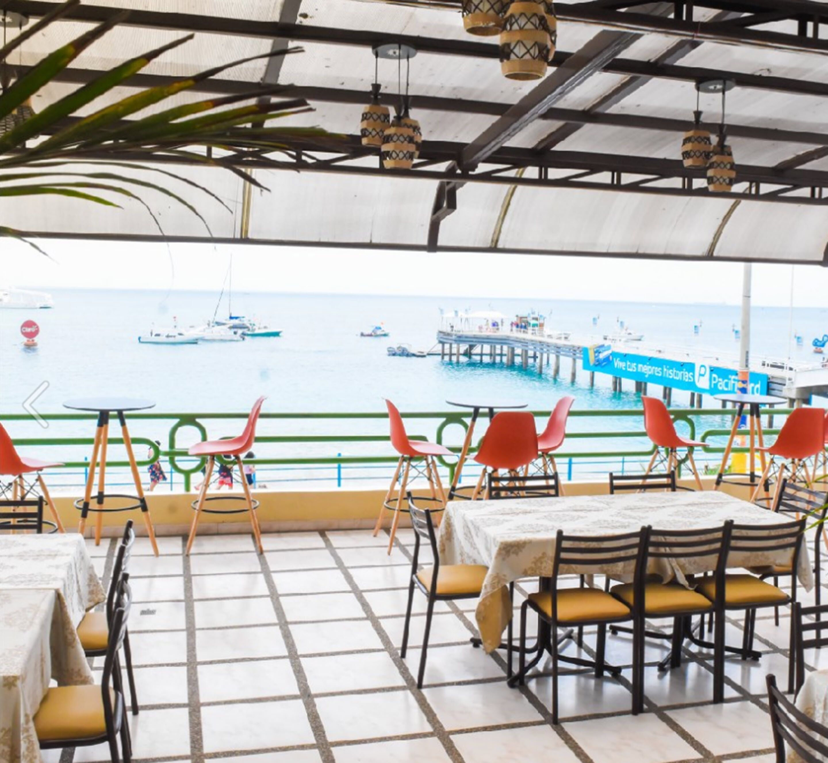 An outdoor dining area with tables and chairs, a view of the sea with boats, and a pier in the background.