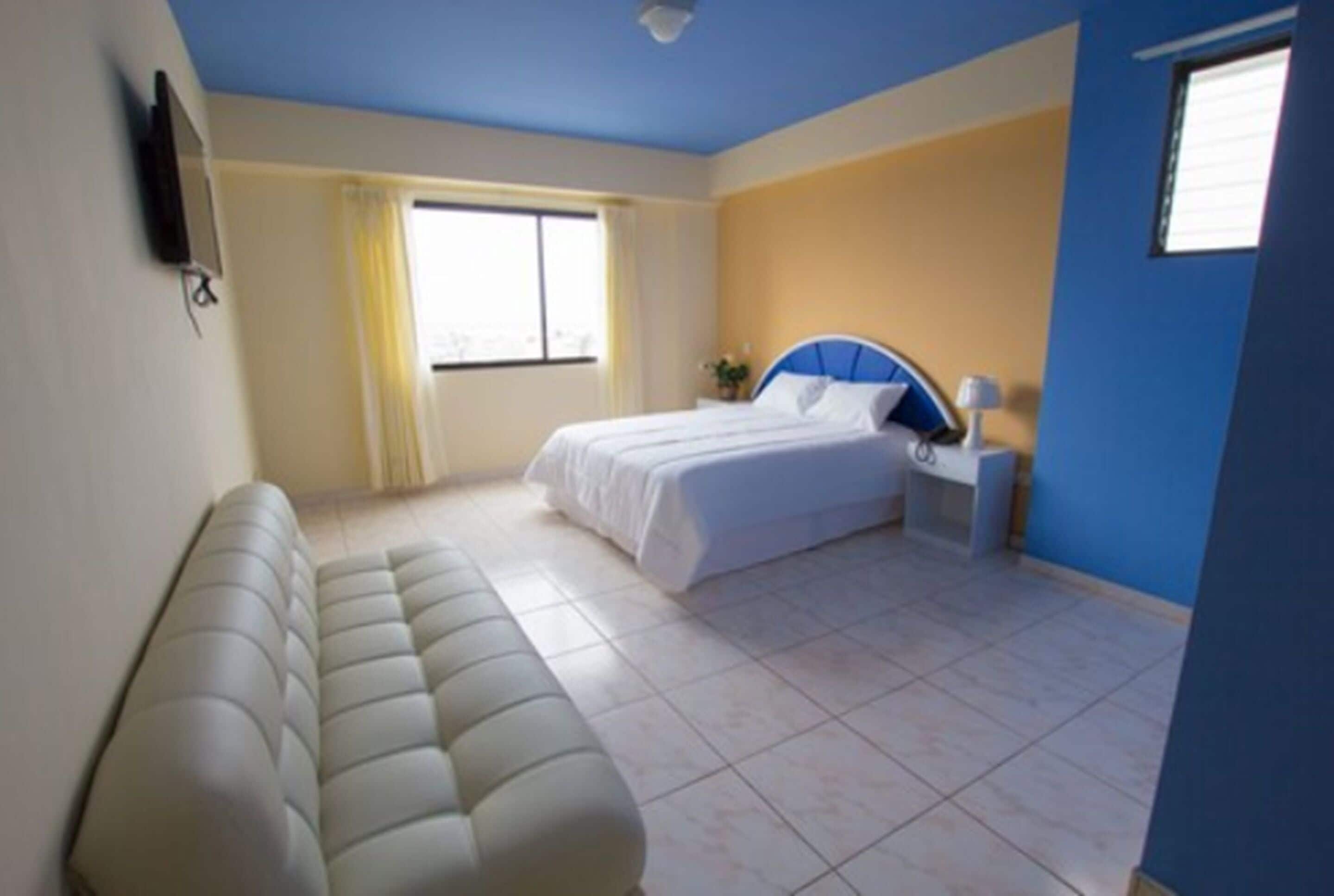 Un dormitorio con cama, mesita de noche, lámpara, sofá y ventana con cortinas.