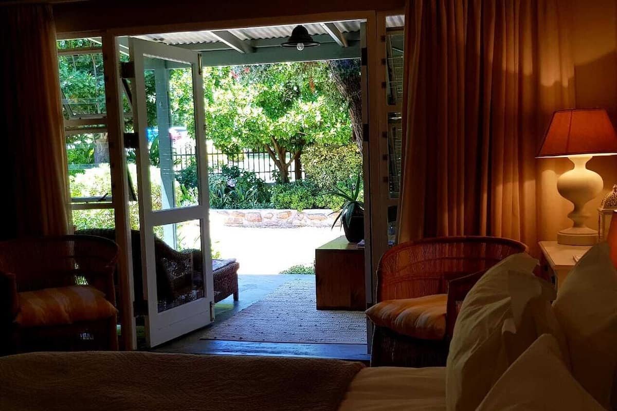 A hotel room with a bed, two chairs, a table, and a view of a garden through an open door.