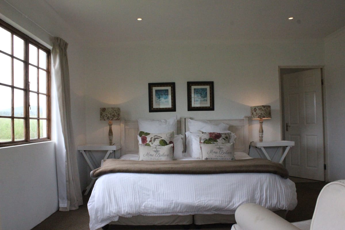 A bedroom with a large bed, two bedside tables, a window with curtains, and two framed pictures on the wall.