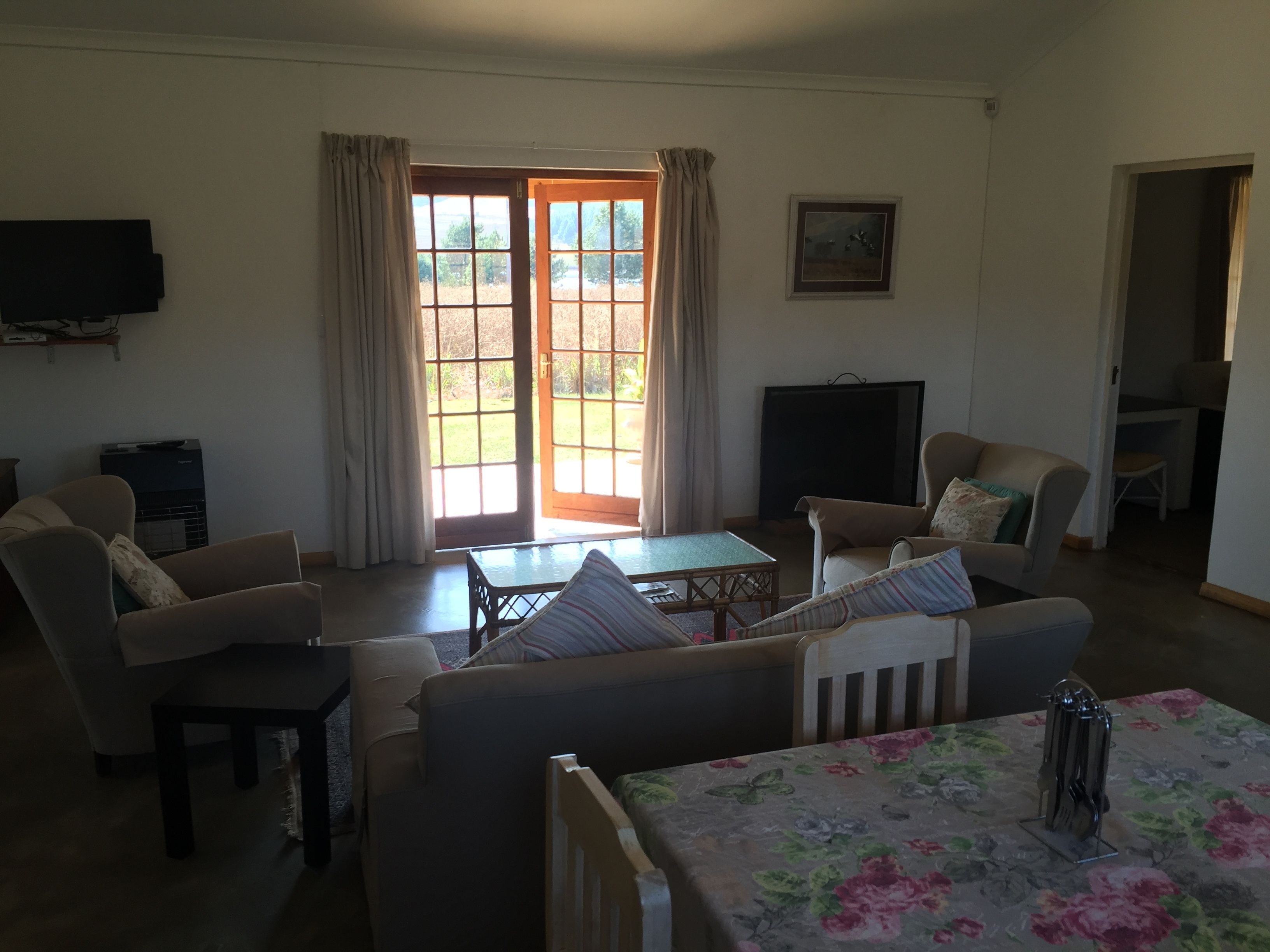 A living room with a sofa, armchairs, a table, and a television. There is a large window with curtains, and a door leading outside.