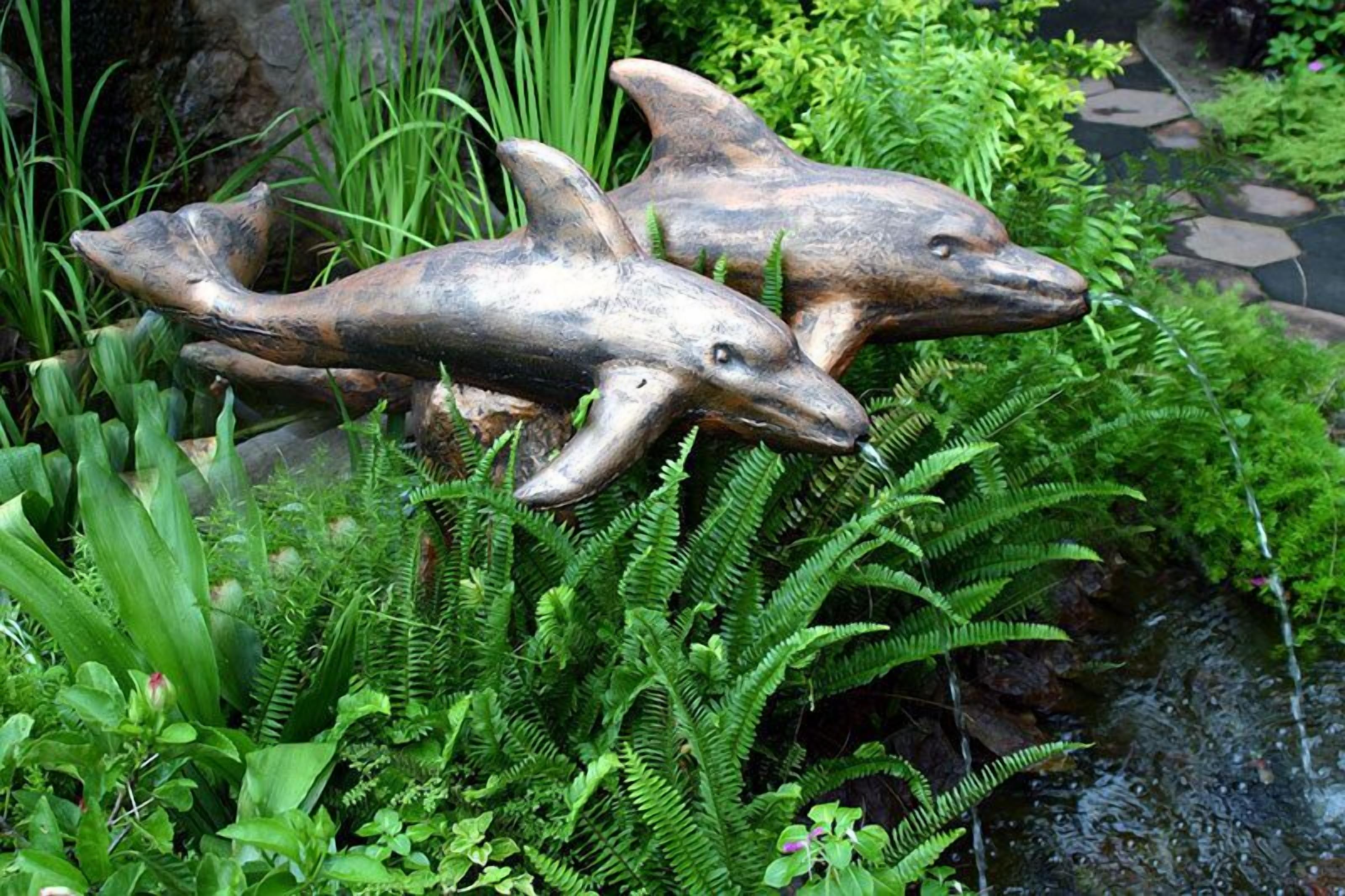 Two dolphin sculptures in a garden with lush green ferns and a small stream.