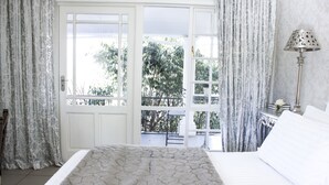 A bedroom with a bed, curtains, a nightstand, a lamp, and a view of trees through a window. - La Riviera Guesthouse (Mohokare)