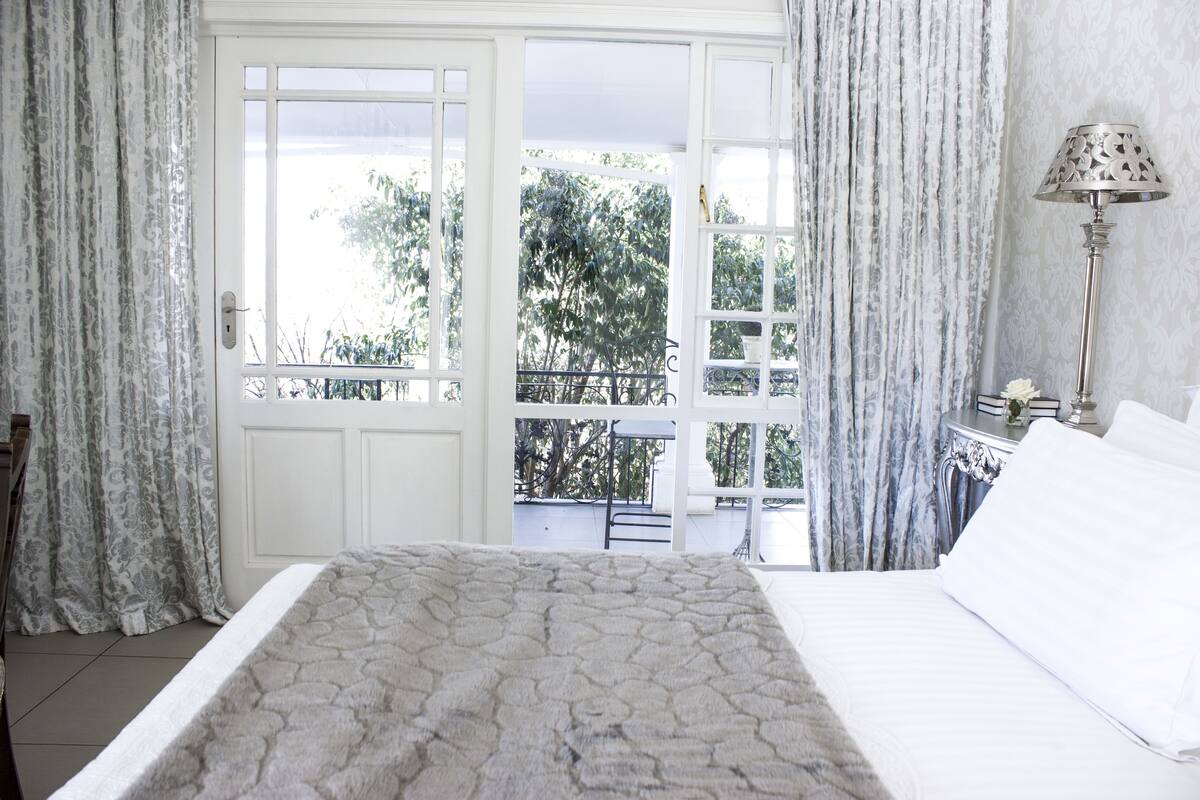 Une chambre à coucher avec un lit, des rideaux, une table de chevet, une lampe et une vue sur les arbres par la fenêtre.