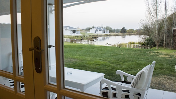 A view from inside a room with a glass door, looking out to a grassy area with a pond, white buildings, and a wooden fence. - La Riviera Guesthouse (Mohokare)