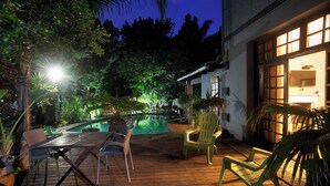 Terraza de madera con piscina, sillas y una casa con ventanas iluminadas.