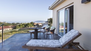A balcony with a table and chairs, a cushioned lounge chair, and a view of the sea and houses. - Lala Panzi B&B (Plettenberg Bay)