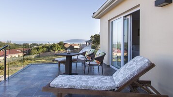 A balcony with a table and chairs, a cushioned lounge chair, and a view of the sea and houses.