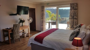 A hotel room with a bed, a desk with a computer, a TV, a fan, and a balcony with a table and chairs. - Lala Panzi B&B (Plettenberg Bay)