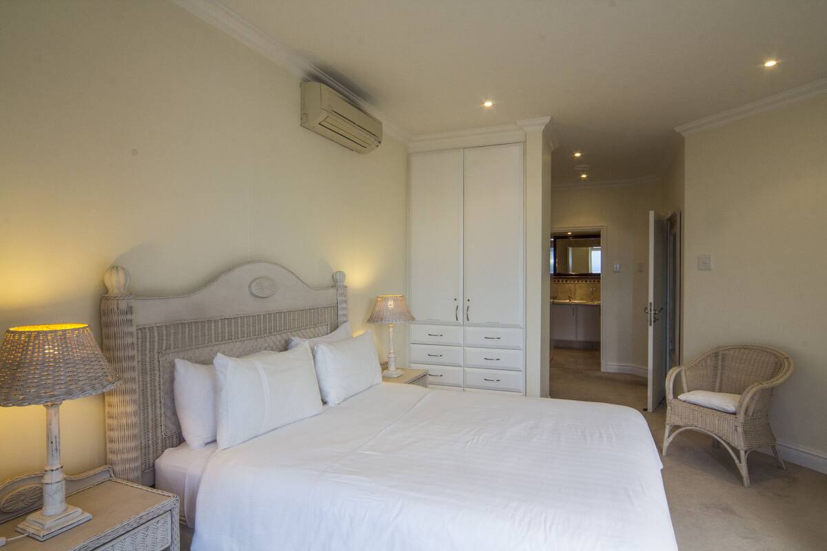 A bedroom with a large bed, a nightstand with a lamp, a wicker chair, and a wardrobe.