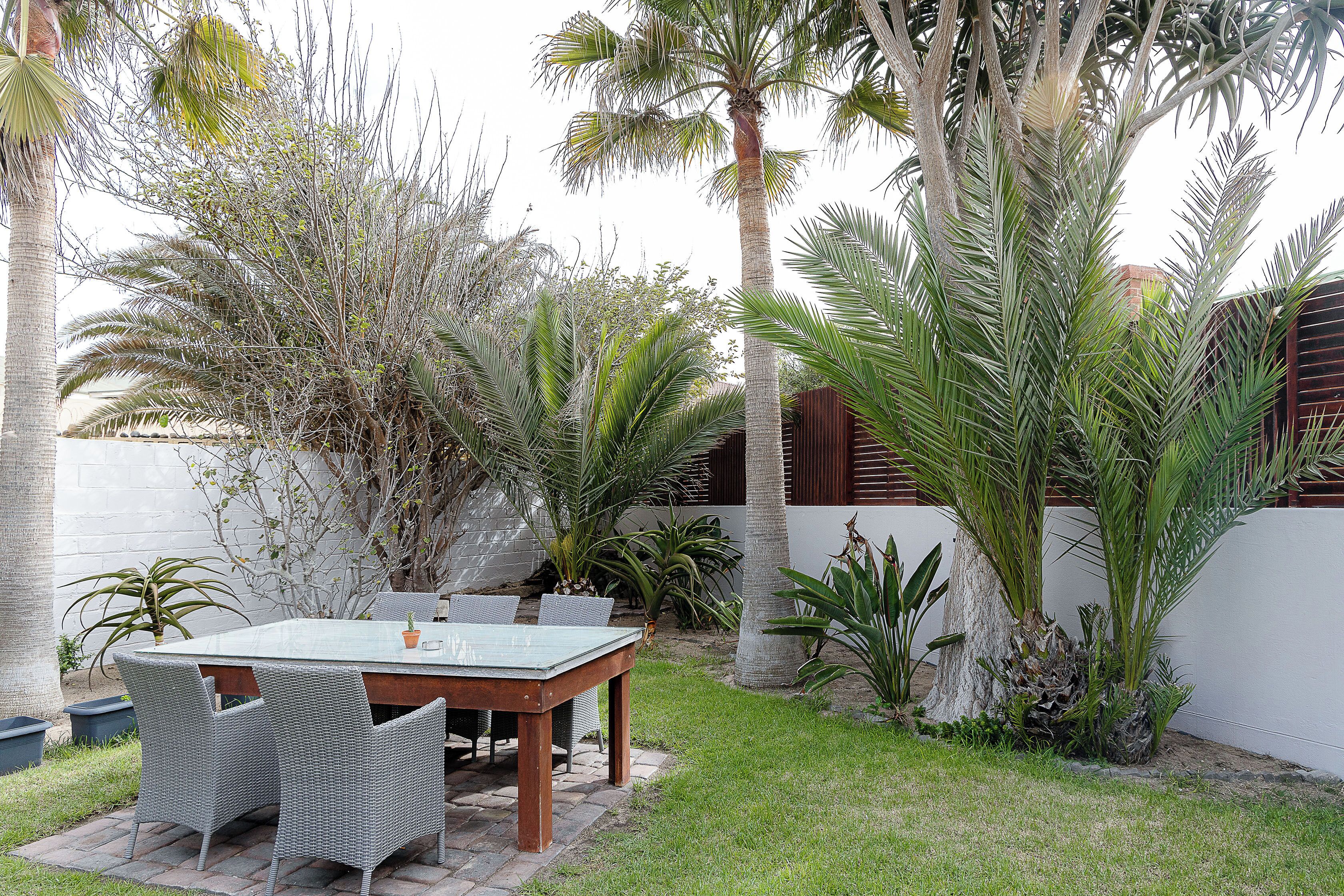 An outdoor patio with a table and chairs, surrounded by various types of plants and trees.