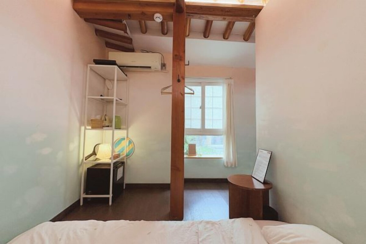 A bedroom with a bed, a wooden ceiling, a window, a shelf with items, and a small table.