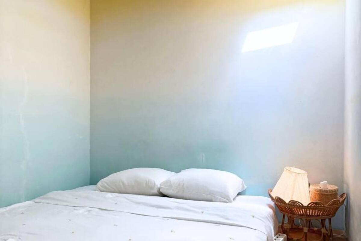 Une pièce avec un lit, des oreillers, une lampe et une chaise en osier.