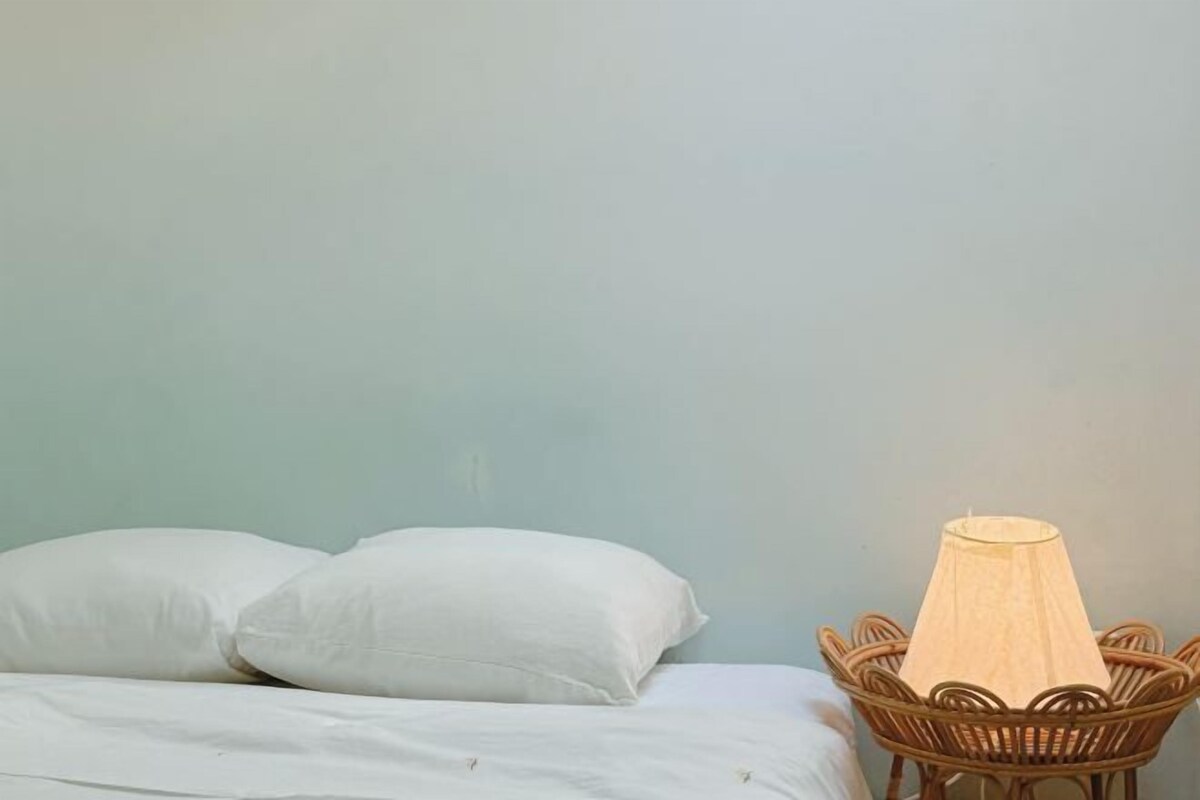 A bed with white bedding and pillows, a wicker chair with a light-colored lampshade, and a plain wall.