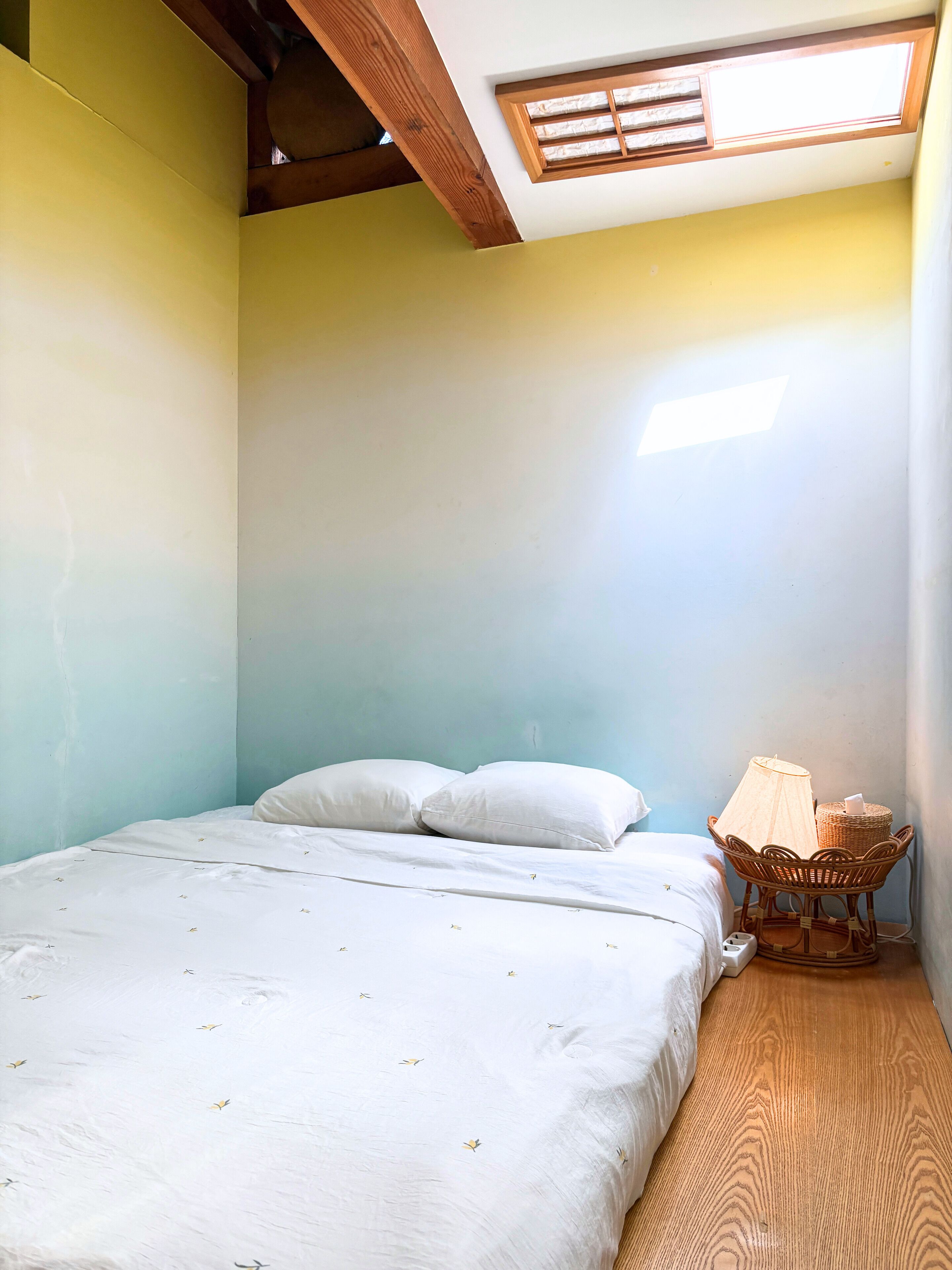 Ein Schlafzimmer mit einem Bett, Kissen, einem Korbstuhl, einem kleinen Tisch und einem Dachfenster.