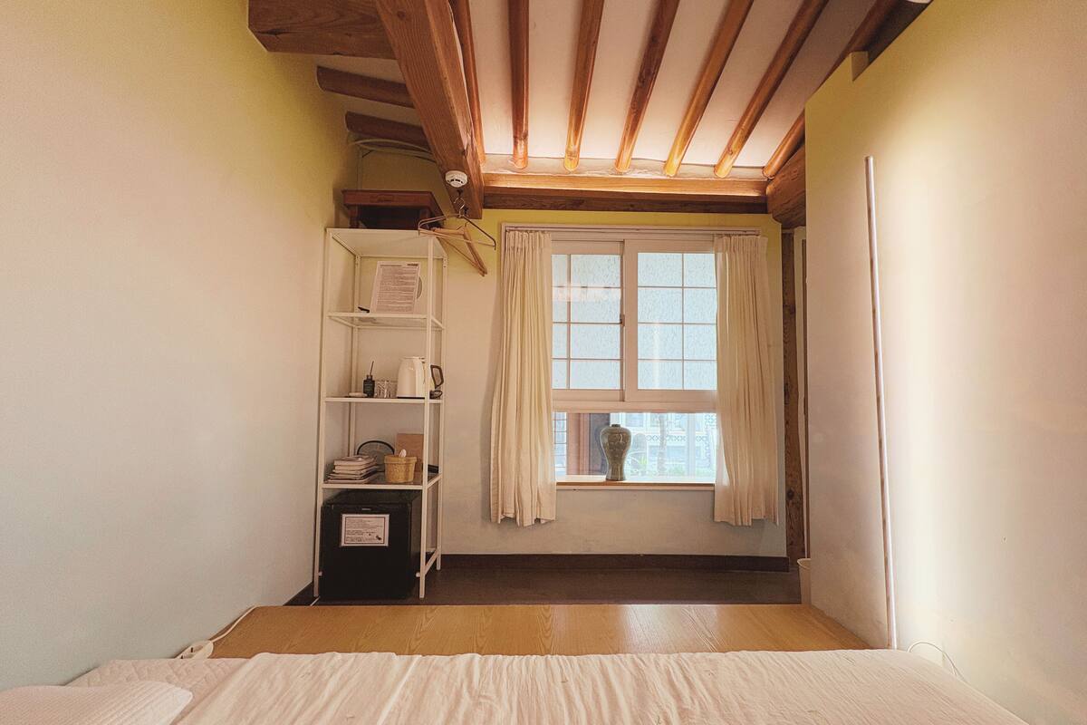 Une petite pièce accueillante, avec un plafond en pente, une fenêtre avec des rideaux, un lit et une étagère où sont rangés divers objets.