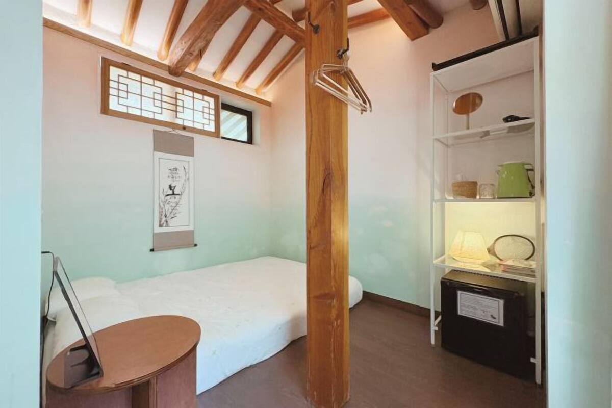 Une chambre avec un lit simple, un plafond avec des poutres apparentes, une étagère contenant des ustensiles de cuisine et un tableau encadré au mur.