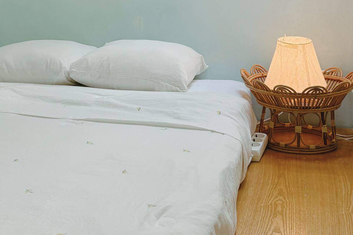 Un lit avec une literie blanche, une table de chevet en osier avec une lampe, et une petite fenêtre au-dessus du lit.