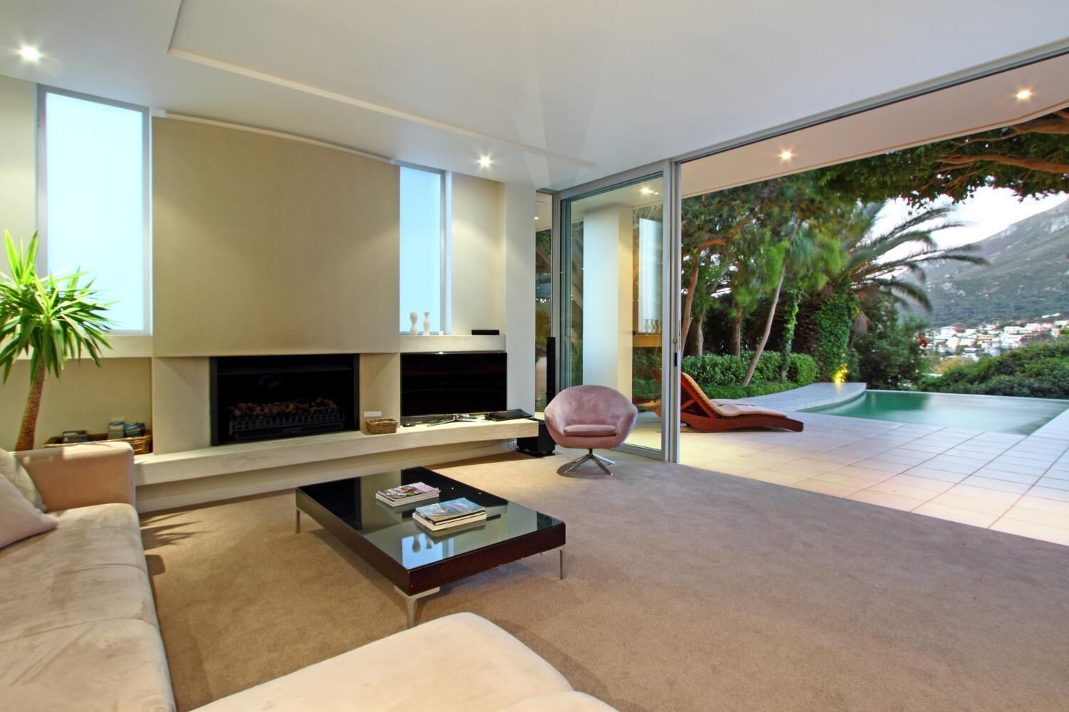 A modern living room with a fireplace, a glass coffee table, a sofa, and a sliding glass door leading to an outdoor area with a pool and greenery.