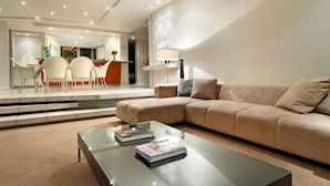 Ein modernes Wohnzimmer mit einem beigen Sofa, einem Glastisch mit Zeitschriften und einem Essbereich mit weißen Stühlen und einem Tisch.