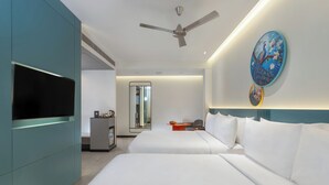 Une chambre d’hôtel moderne équipée d’un téléviseur à écran plat, d’un ventilateur de plafond, de deux lits avec une literie blanche, d’une petite table sur laquelle se trouve un panier de fruits et d’une œuvre d’art décorative accrochée au mur.