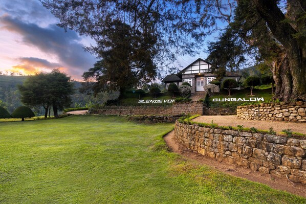A bungalow with a stone retaining wall, surrounded by a grassy lawn and trees. - Scottish Planter Glendevon Bungalow (Walapane)