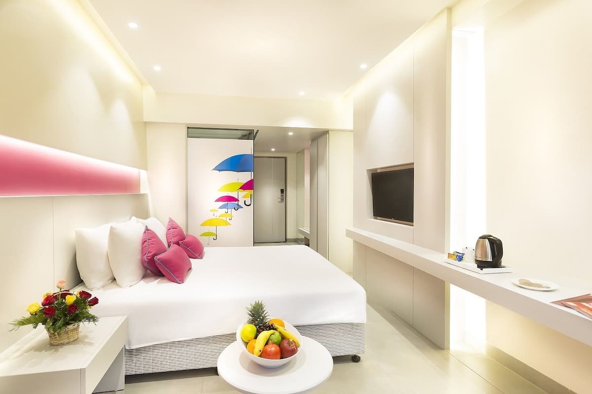 A modern hotel room with a large bed, a bedside table with a fruit bowl, a TV, and a desk with a kettle.