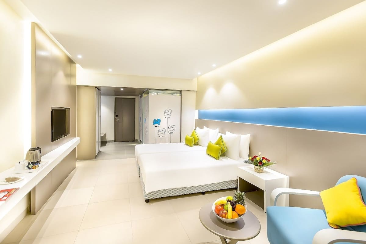 A modern hotel room with a large bed, bedside table, and a small round table with fruit.