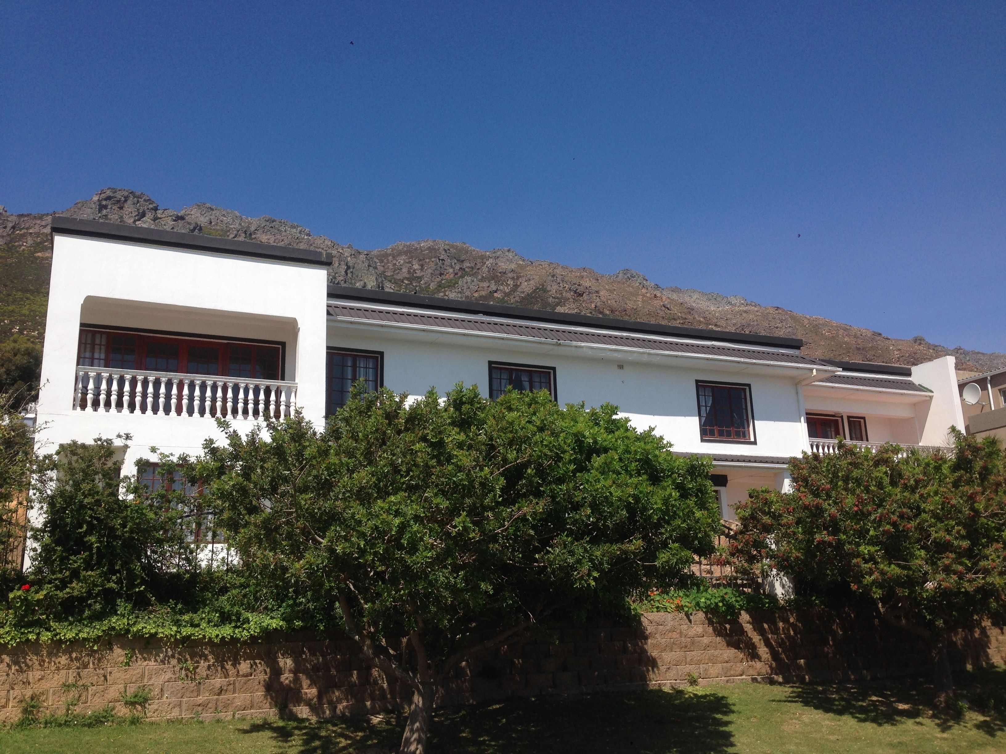 A two-story house with a balcony, surrounded by greenery and mountains in the background.