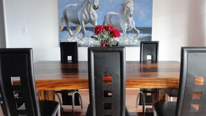 A dining area with a wooden table and black chairs, a painting of horses on the wall, and a vase with red flowers. - Helderberg Guesthouse (Cape Town)