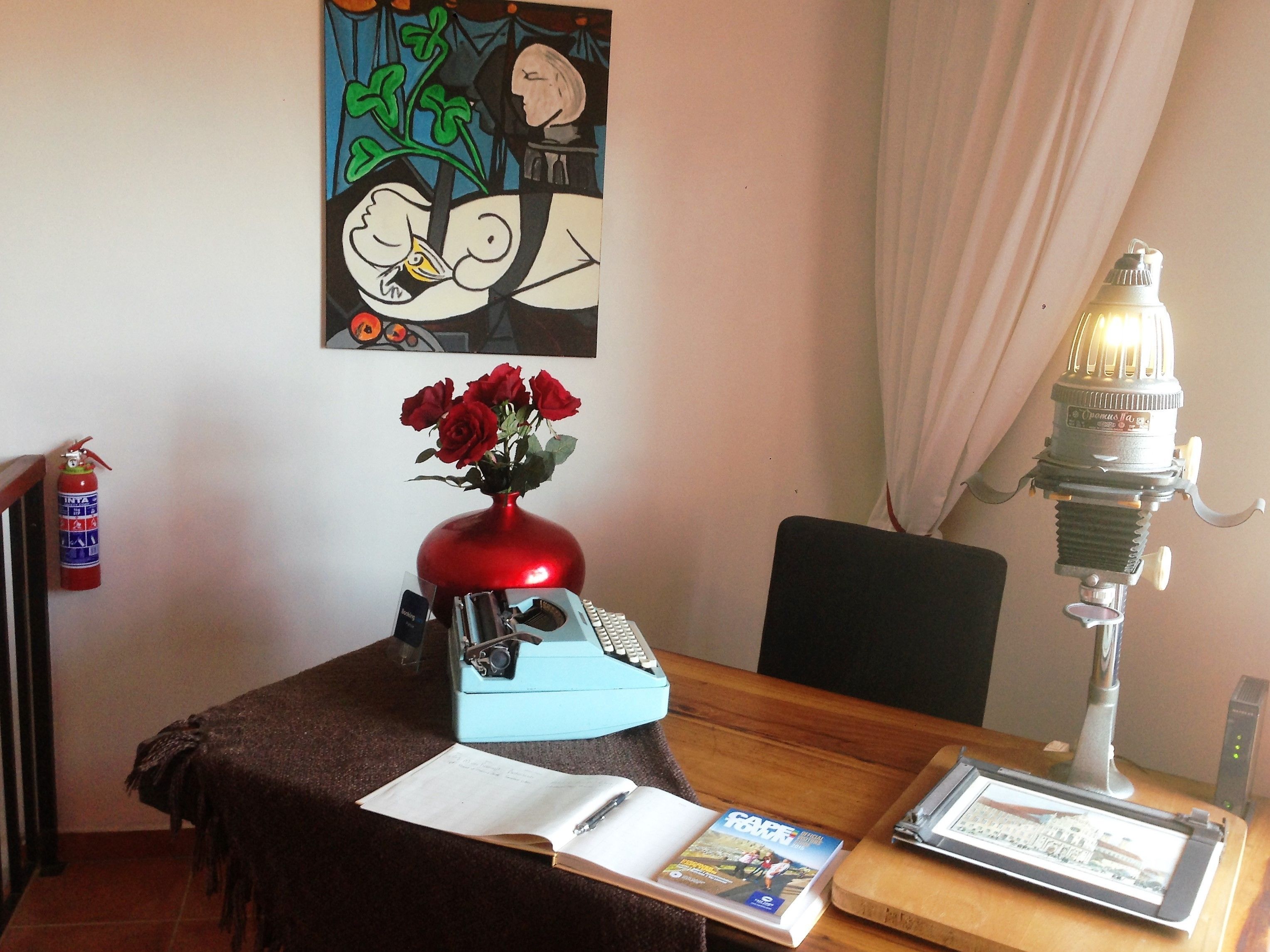 Una habitación con un escritorio, una máquina de escribir antigua, un jarrón con rosas rojas, un extintor, una lámpara y un cuadro en la pared.