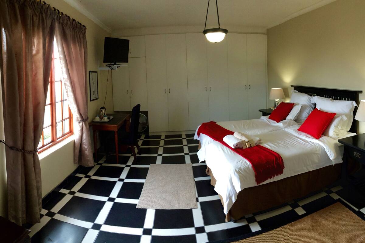 Une chambre d’hôtel avec un sol à carreaux, un lit avec une literie rouge et blanche, un bureau avec une chaise, un téléviseur fixé au mur et un grand miroir.