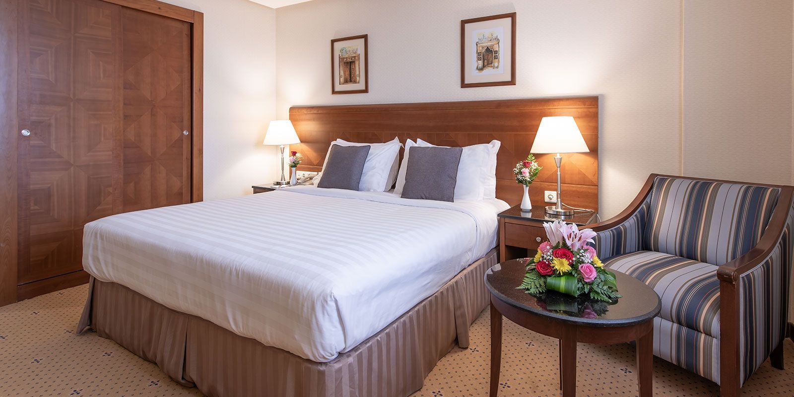 Ein Hotelzimmer mit einem großen Bett, zwei Nachttischen mit Lampen, einem Sessel und einem kleinen Tisch mit Blumen.