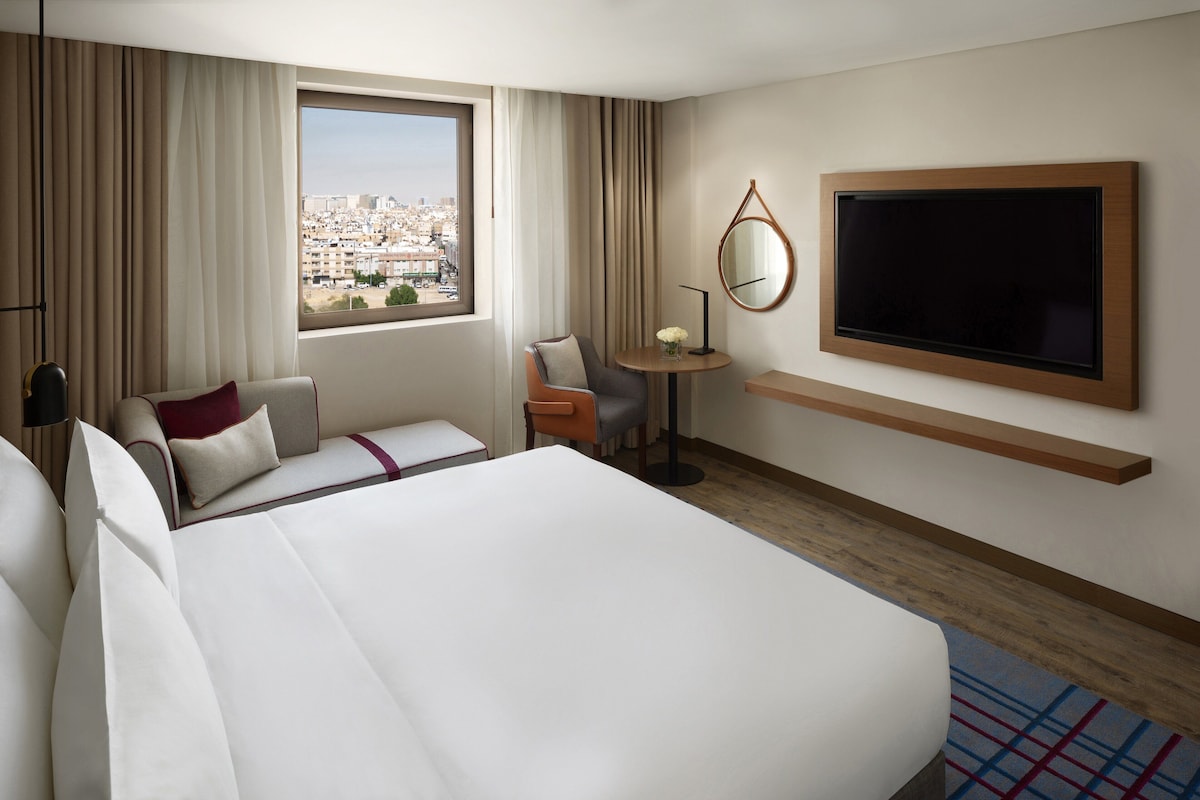 A hotel room with a large bed, a desk, a chair, a TV, and a view of the city.