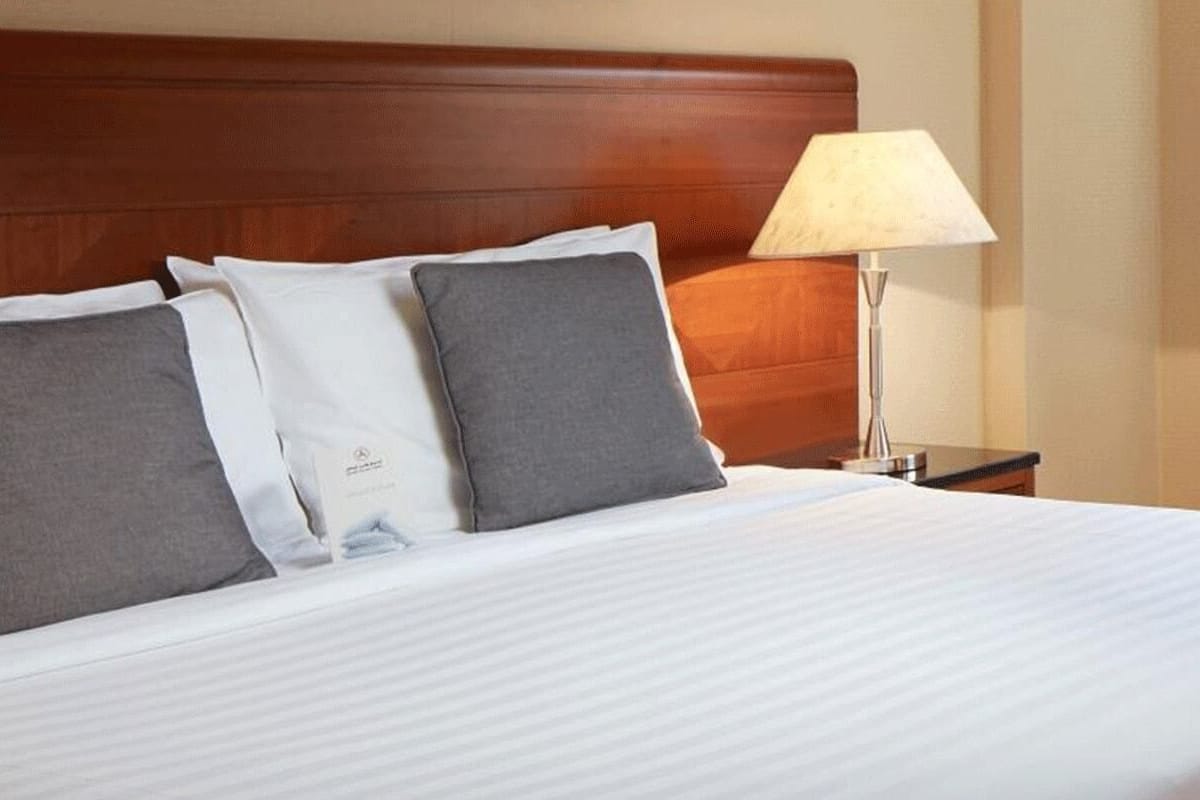 Un lit bien fait, avec des draps blancs et deux oreillers gris, une tête de lit en bois et une lampe de chevet.