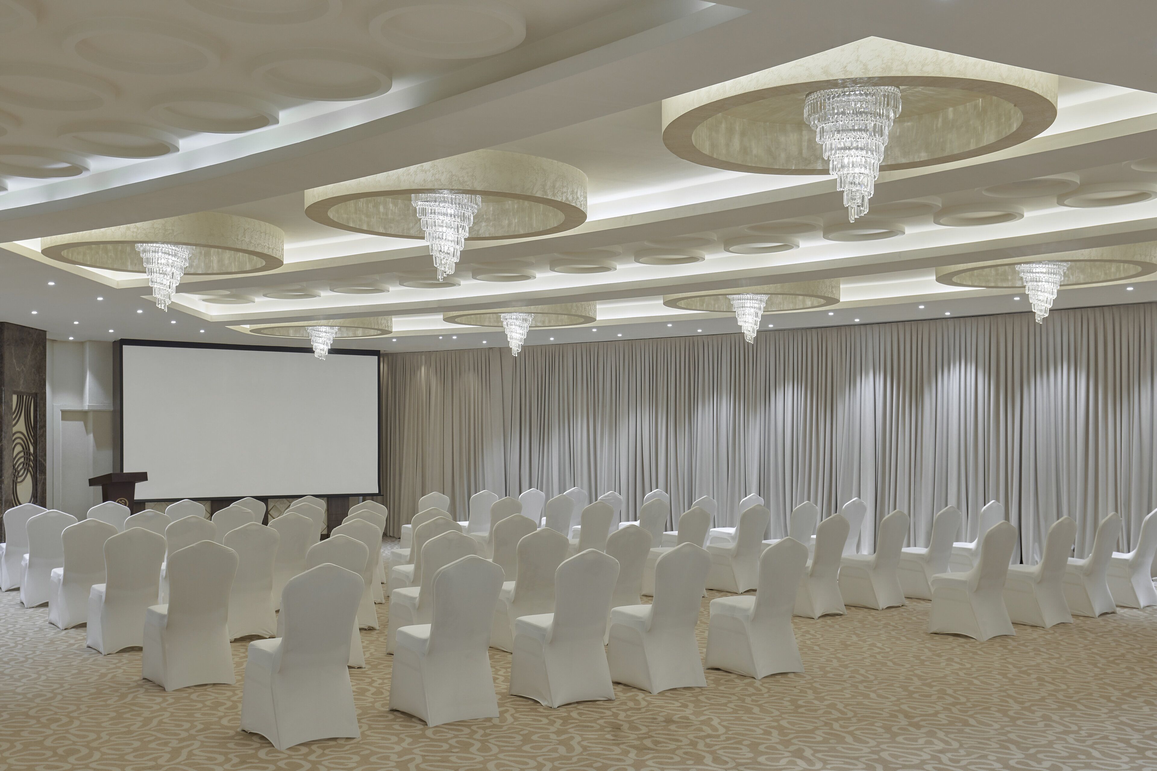 Un salón de conferencias con filas de sillas blancas, una pantalla grande y candelabros ornamentales en el techo.