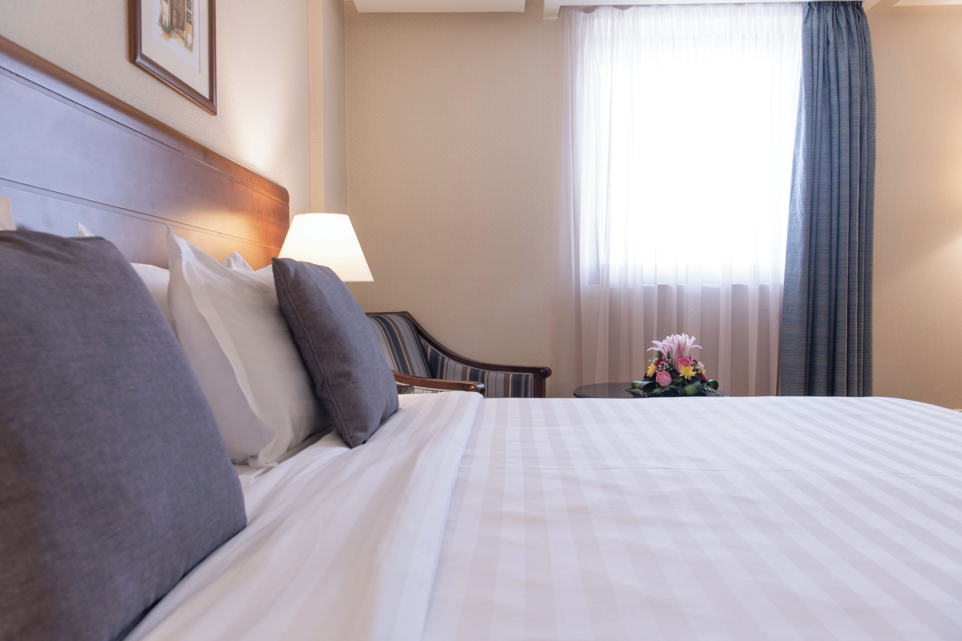 A neatly made bed with white and gray linens, a wooden headboard, a bedside table with a lamp, and a window with sheer curtains.