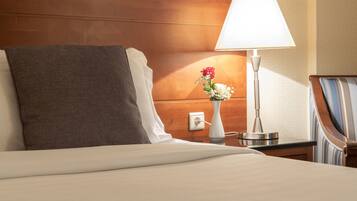 A neatly made bed with a gray pillow, a white lamp, and a vase with flowers on a wooden nightstand.