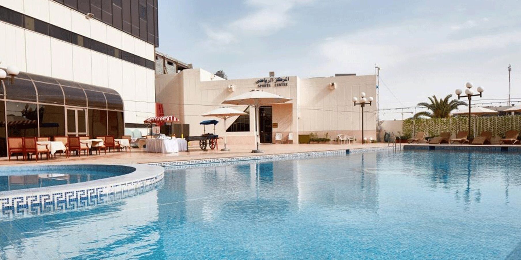 Una piscina d'hotel amb gandules i ombrelles, un centre esportiu i un edifici amb grans finestres.