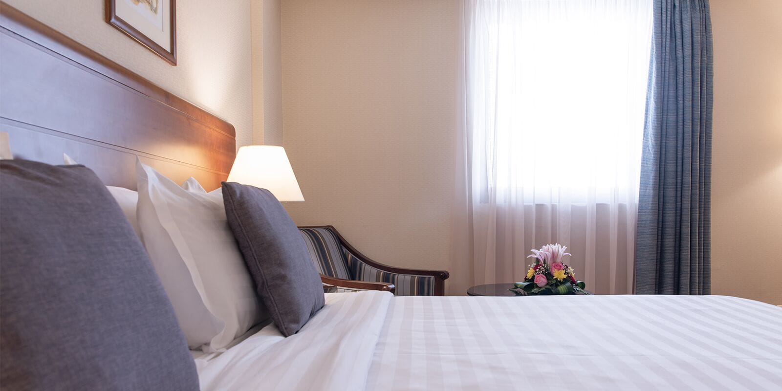 A hotel room with a neatly made bed, a bedside table with a lamp, a chair, and a window with curtains.