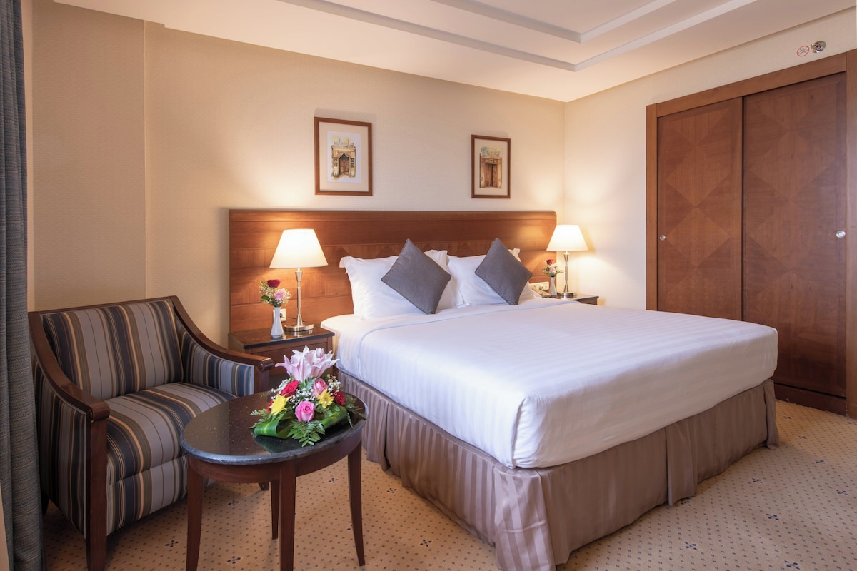 A hotel room with a bed, a sofa, a small table with flowers, two bedside lamps, and a wardrobe.