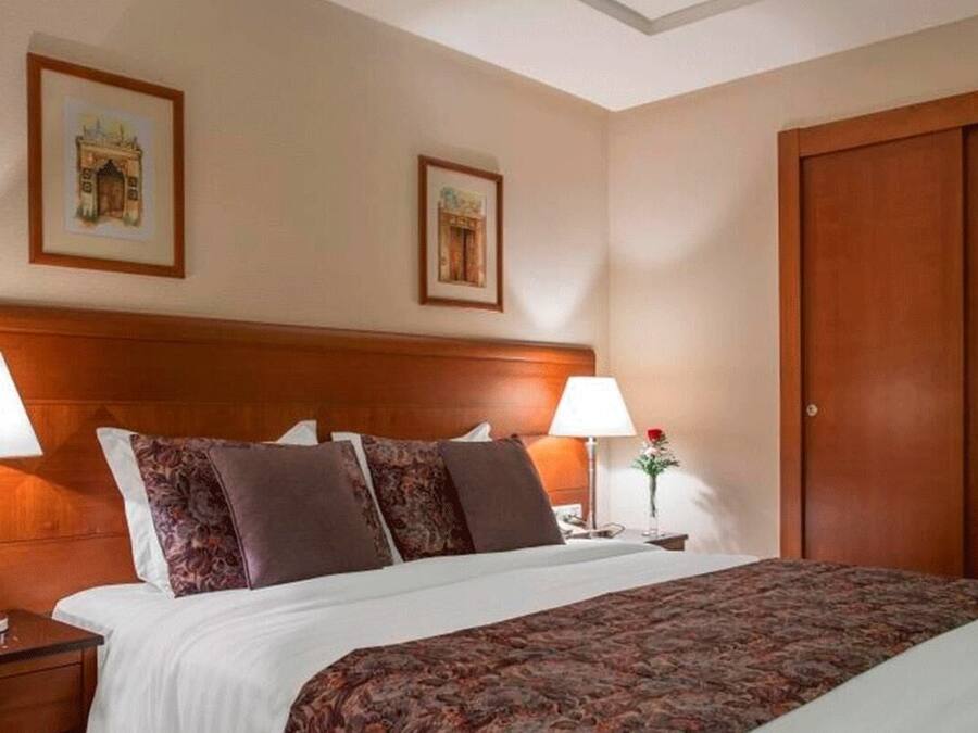 A hotel room with a large bed, two bedside lamps, a nightstand, a wooden headboard, and two framed pictures on the wall.